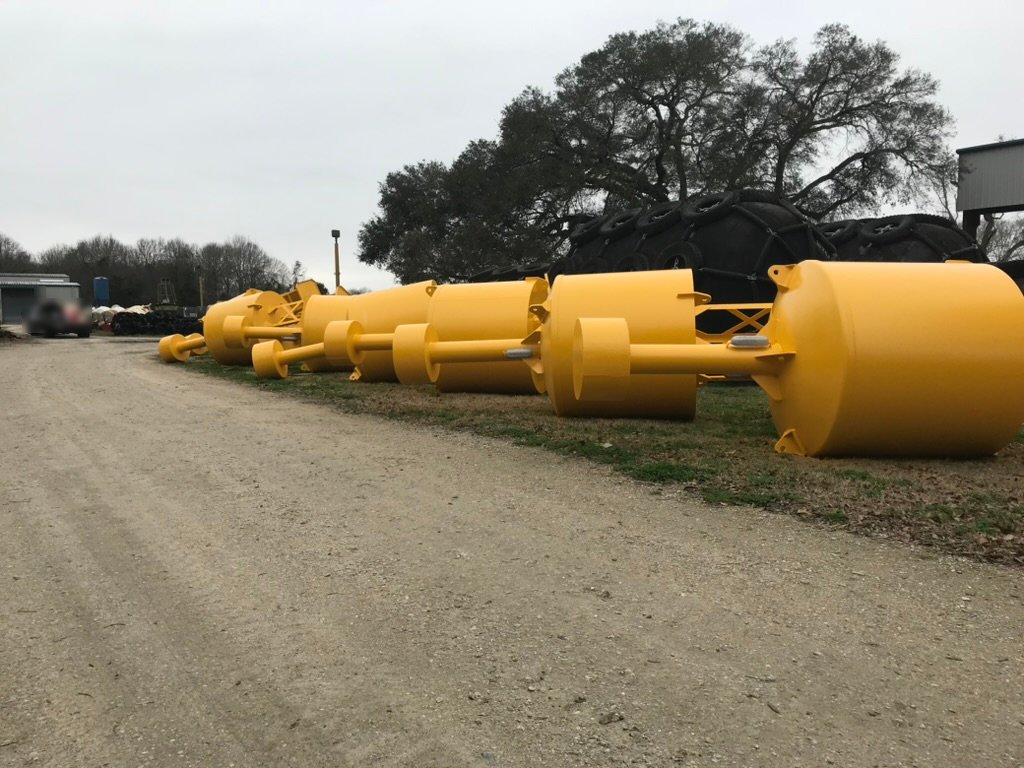 Row of yellow-painted BL 620 steel buoys lying on their sides on the grass in the Wet Tech yard, with black pneumatic fenders and a large oak tree behind them