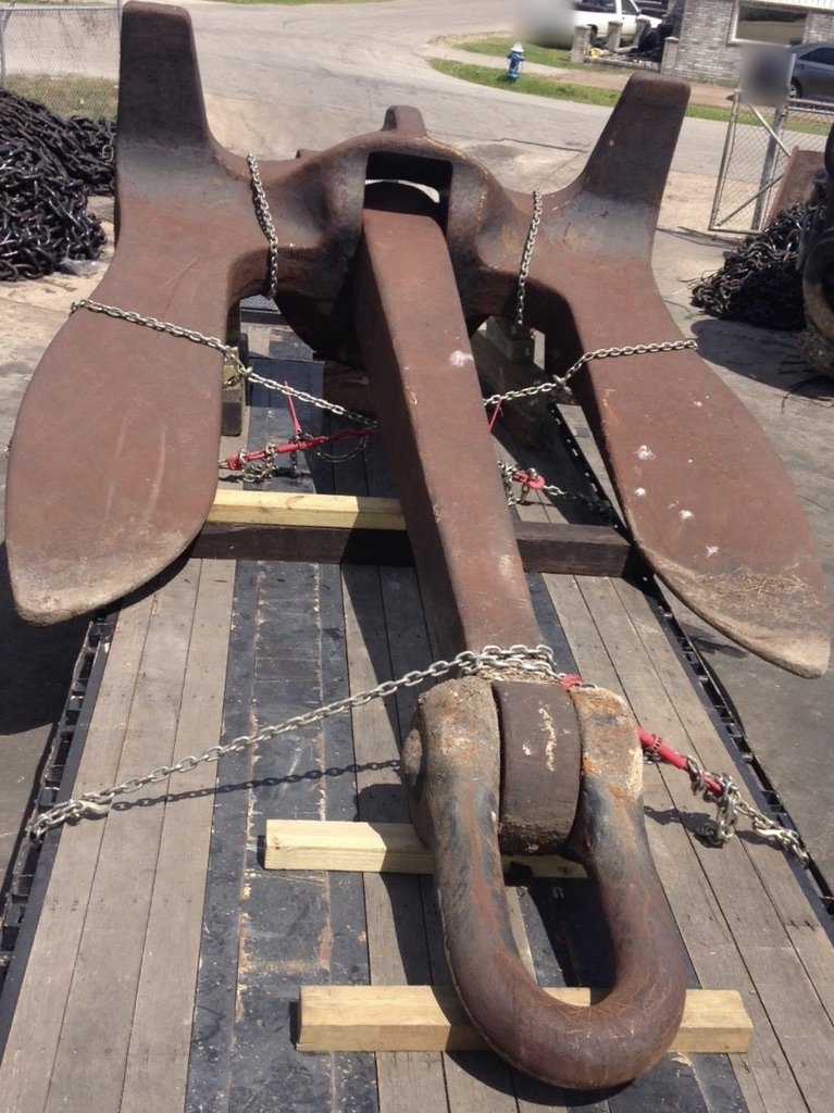 Large rusty fluke anchor chained down on wooden blocks on a flatbed trailer, with piles of chain beside it