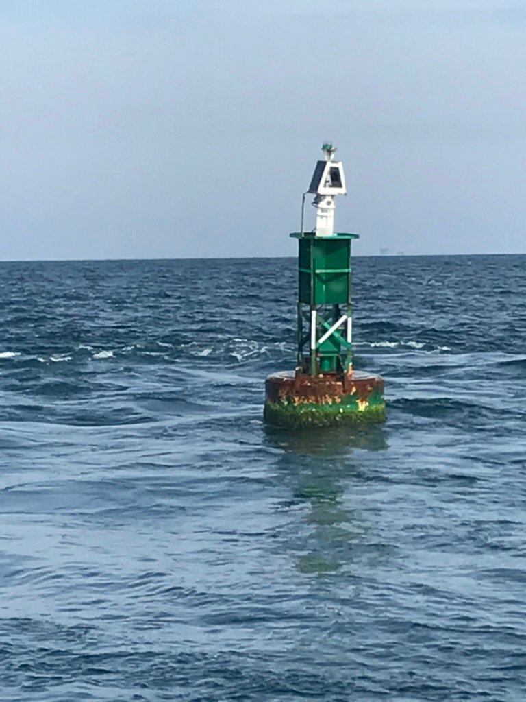 Green steel BL 620 lighted sound buoy on station at sea, with a solar panel and light on top of its tower and marine growth on the round hull